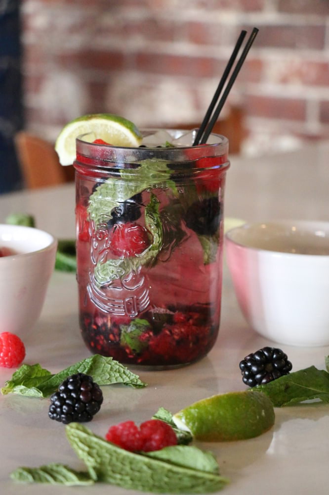 A photo of a raspberry and blackberry mojito on a white counter top with berries, lime wedges, and mint placed around the cup for decoration.